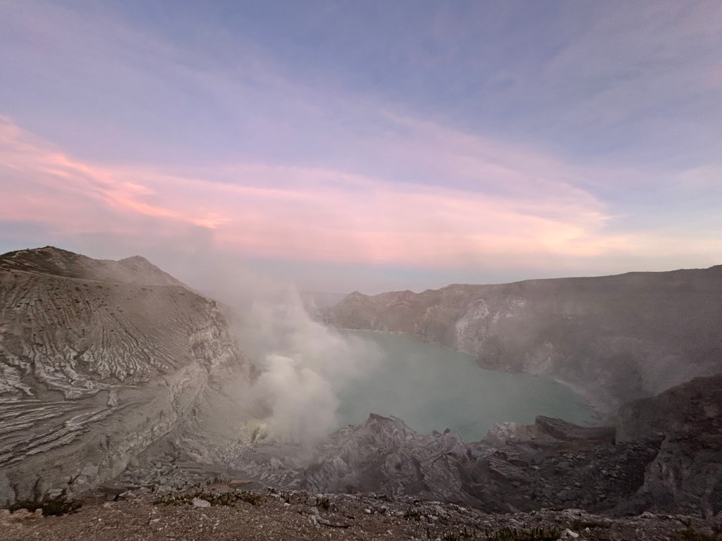 A view of a volcanic crater with a turquoise lake surrounded by steep, rocky cliffs under a colorful sky at dawn.